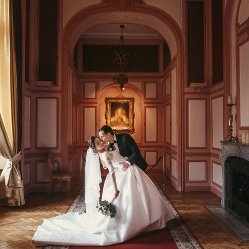 gorgeous bride with bouquet and stylish groom gently kissing in luxury room in hotel. rich wedding couple embracing. romantic sensual moment of newlyweds in amazing classic indoors