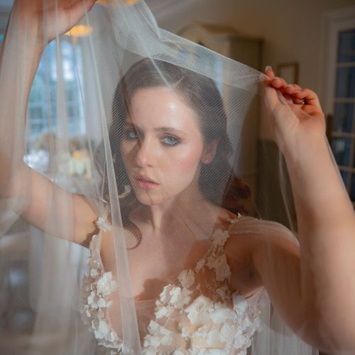 Mariée lumineuse tenant son voile, regard intense et robe florale en relief